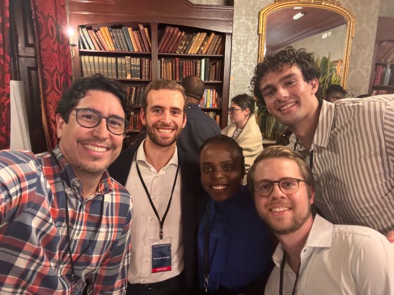 Five smiling attendees wearing event lanyards pose together for a selfie in a book-lined room.